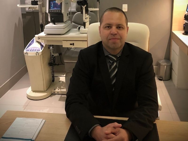 Dr. Flávio Braga da Silva, oftalmologista do Centro Médico Oftalmológico, retrato em consultório com equipamentos de exame oftalmológico ao fundo