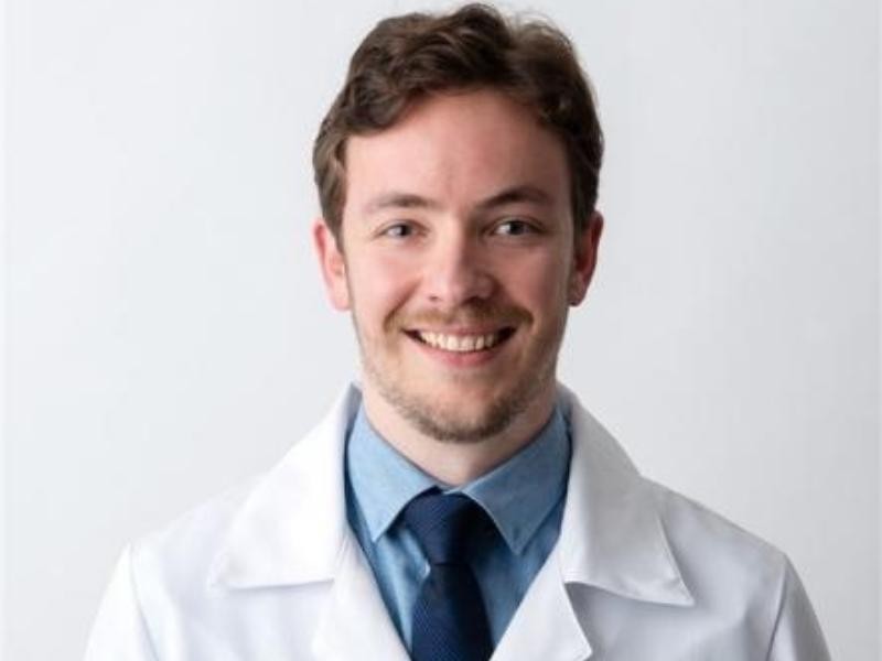 Retrato profissional do Dr. Guilherme Herrmann Matos, médico oftalmologista, jaleco branco, camisa azul e gravata, sorrindo, fundo neutro claro, fotografia realista de estúdio, composição frontal